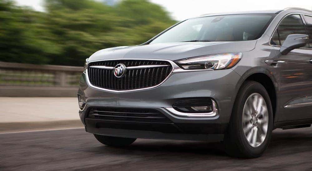 The front half of a gray 2021 Buick Enclave is shown while driving on a tree-lined road.