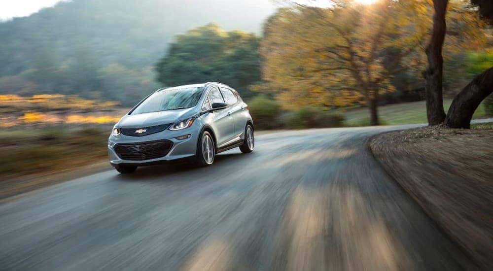 A silver 2018 Chevy Bolt EV is driving on a winding road past trees.