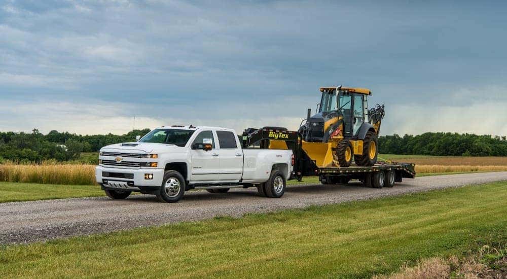 A white 2016 Chevy Silverado 3500HD is towing construction equipment on a gravel road.